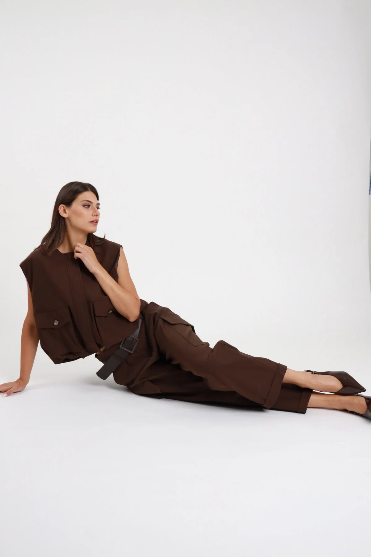 Pantalone Cargo Tasconi Vita Alta marrone cioccolato 78% viscosa vita alta pieghe tasconi cargo gamba barrel ballon risvolto cropped posa seduta laterale pavimento - modella intera seduta laterale pavimento posa mano collo sguardo laterale capelli castani lisci sfondo bianco gilet marrone coordinato cintura ecopelle nera
