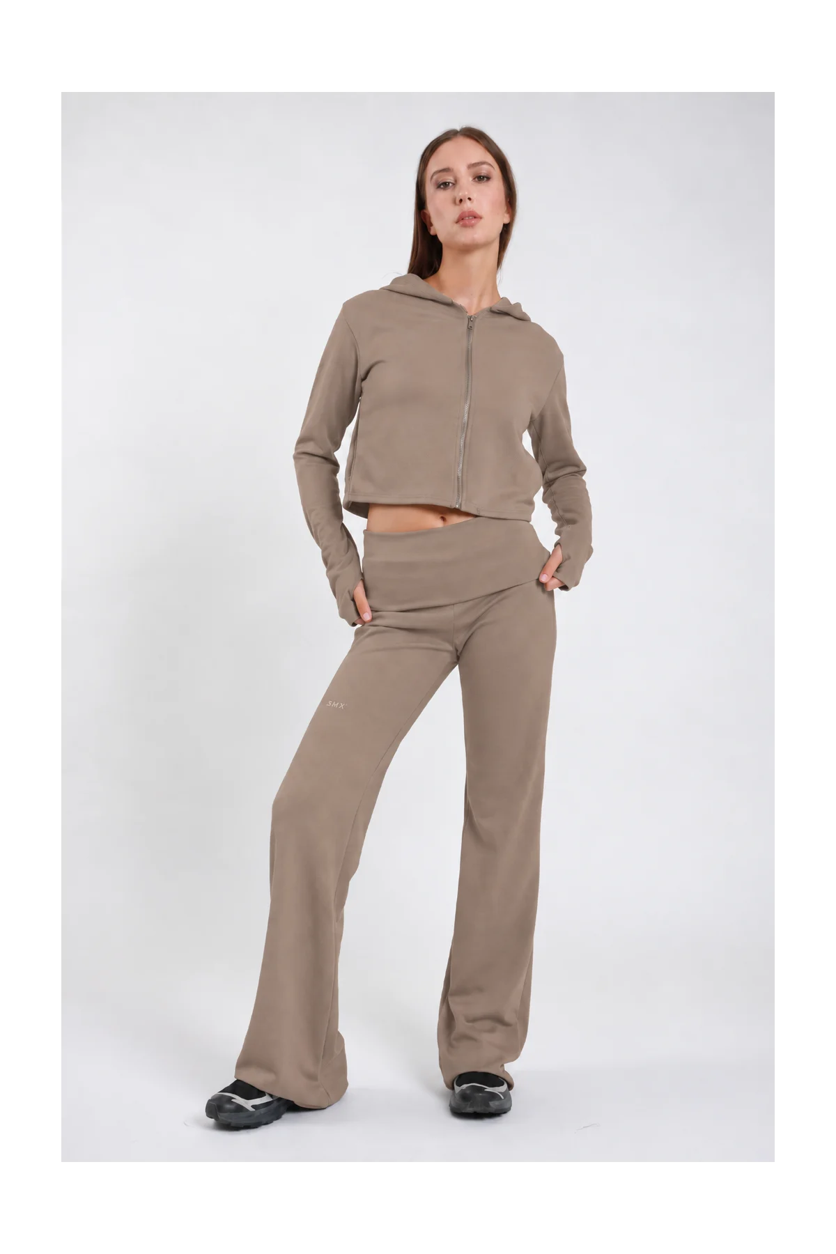 Pantaloni Flare DAYTHRU grigio taupe tortora elastico fold-over vita bassa logo SMX coscia gamba flare bootcut frontale intera 3/4 posa dinamica mano fianco sguardo laterale - modella frontale intera 3/4 posa dinamica mano fianco sguardo laterale capelli castani lisci sfondo bianco felpa crop zip taupe coordinata sneakers nere chunky