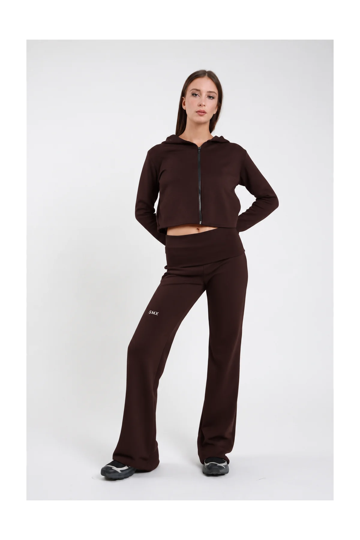 Pantaloni Flare DAYTHRU marrone cioccolato scuro elastico fold-over vita bassa logo SMX bianco coscia ben visibile gamba flare bootcut frontale intera posa statica mano fianco sguardo frontale - modella frontale intera posa statica mano fianco sguardo frontale capelli castani lisci sfondo bianco felpa crop zip marrone coordinata sneakers nere