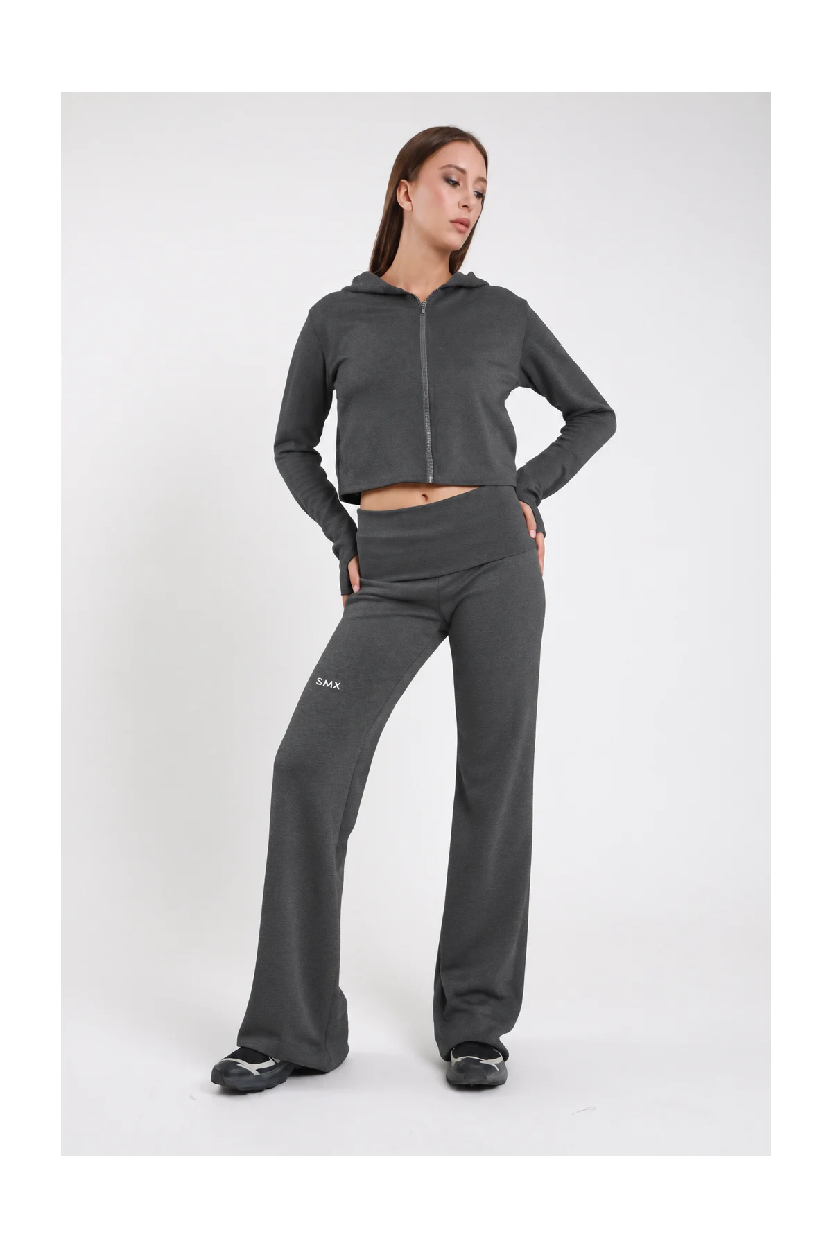Pantaloni Flare DAYTHRU grigio antracite elastico fold-over vita bassa logo SMX bianco coscia ben visibile gamba flare bootcut frontale intera 3/4 posa dinamica mano fianco sguardo basso - modella frontale intera 3/4 posa dinamica mano fianco sguardo basso capelli castani lisci sfondo bianco felpa crop zip antracite coordinata sneakers chunky bianche grigie