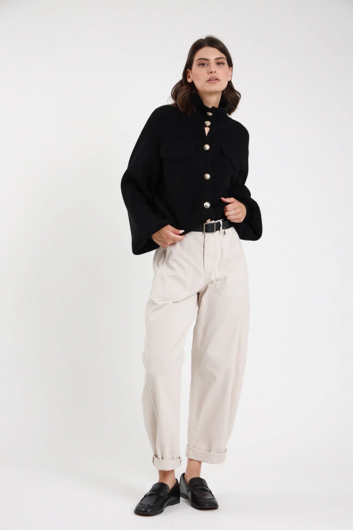 Pantalone Stellina ovetto balloon vita alta pinces risvolto affusolato marrone scuro terra frontale intera - modella frontale intera mani fianchi sguardo laterale capelli castani scuri sfondo bianco maglia manica corta nera cintura nera décolleté neri kitten heel