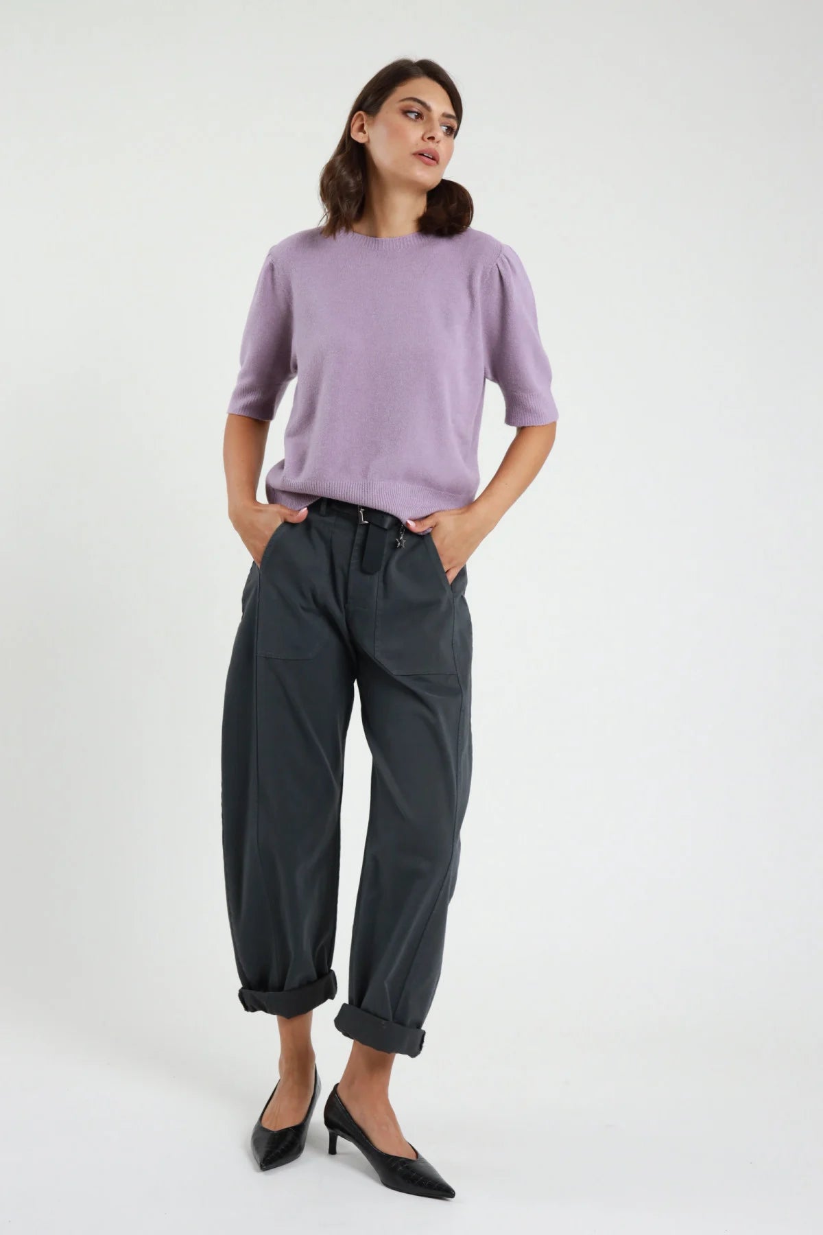 Pantalone Stellina ovetto balloon vita alta pinces risvolto affusolato bordeaux prugna frontale intera - modella frontale intera mani fianchi sguardo laterale capelli castani scuri sfondo bianco maglia manica corta grigia cintura nera mule tacco bordeaux kitten heel