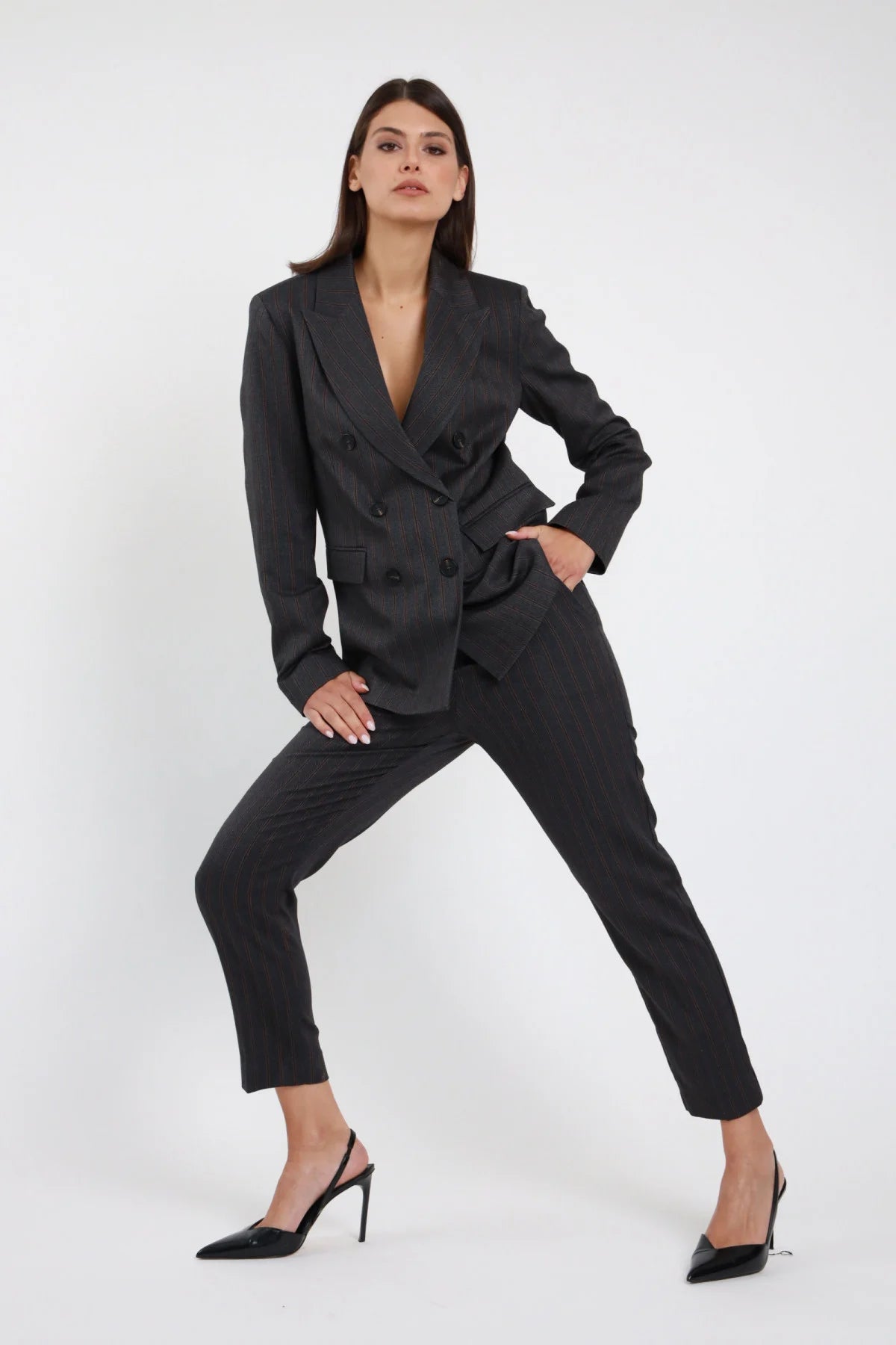 Pantalone slim gessato Susy Mix Pre-Fall grigio antracite pinstripe bordeaux vita alta gamba slim sigaretta affusolata lunghezza caviglia frontale intera mano fianco posa dinamica - modella intera frontale mano fianco posa dinamica capelli scuri lisci sfondo bianco blazer doppiopetto gessato coordinato tailleur slingback neri abbinati