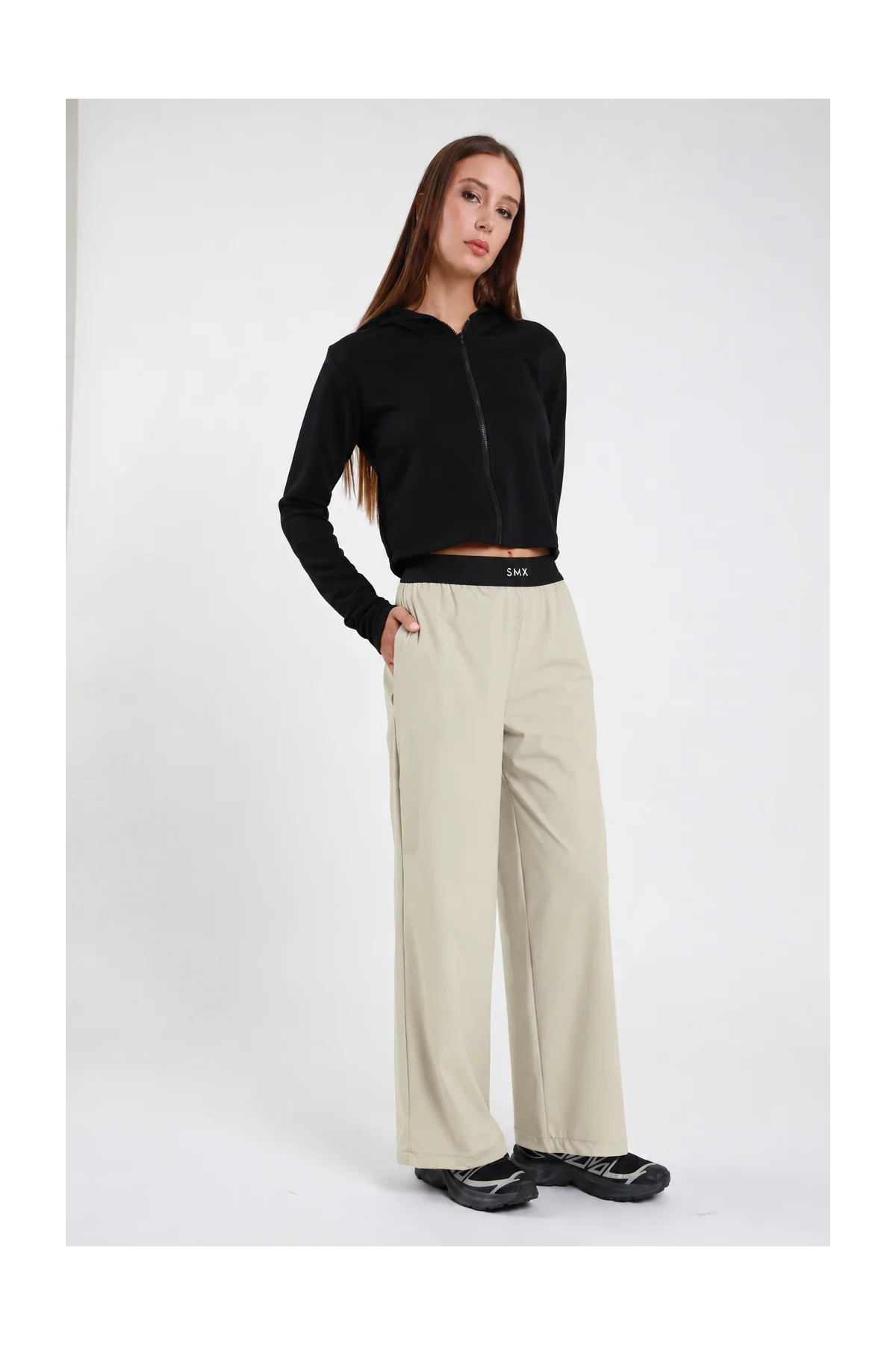 Pantaloni Wide-Leg Soft Air DAYTHRU sabbia beige chiaro elastico nero logo SMX bianco gamba wide piega frontale tasche laterali silhouette palazzo lunga frontale intera 3/4 posa statica mano tasca sguardo frontale - modella frontale intera 3/4 posa statica mano tasca sguardo frontale capelli castani lisci sfondo bianco felpa zip crop nera sneakers nere chunky