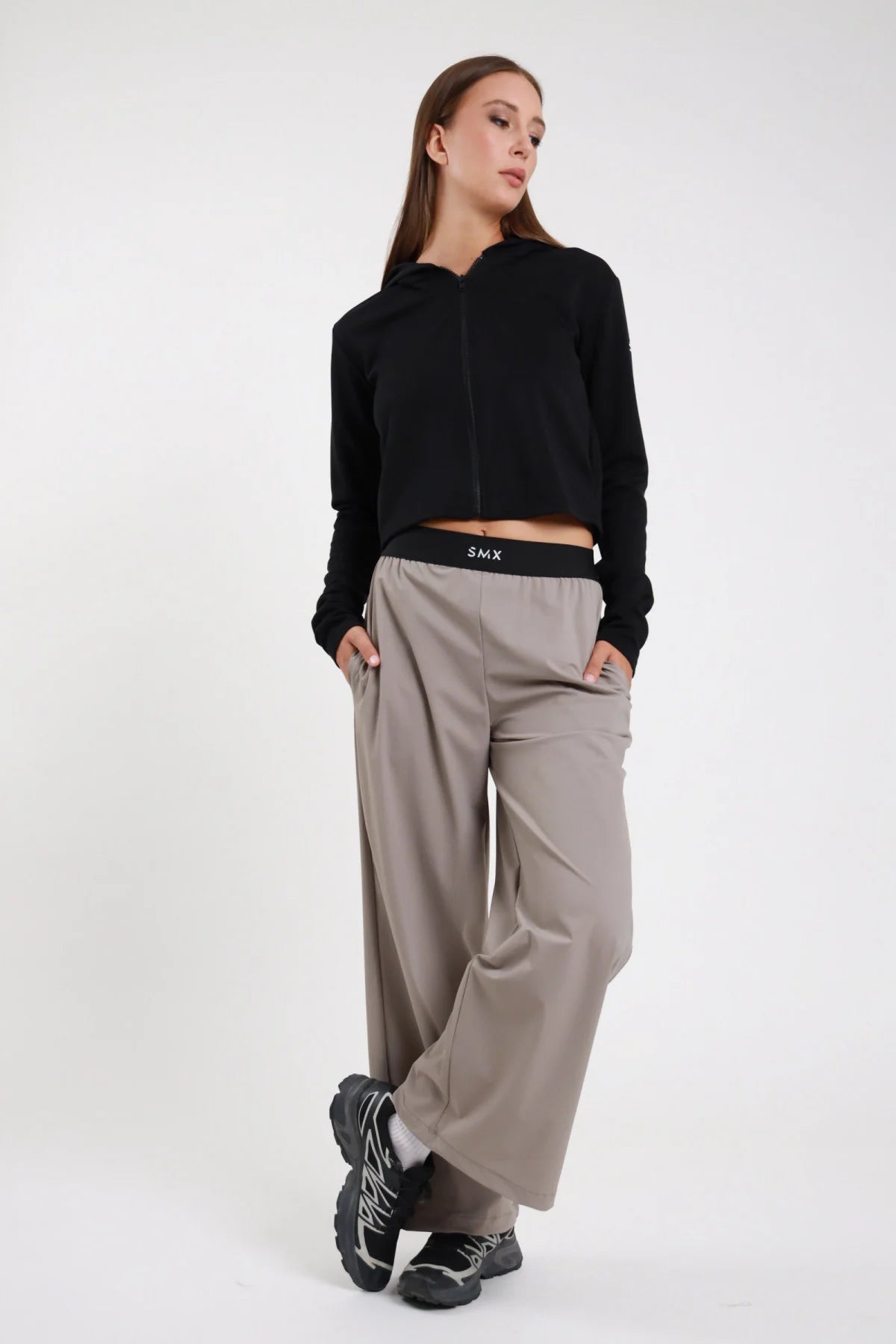 Pantaloni Wide-Leg Soft Air DAYTHRU grigio taupe tortora elastico nero logo SMX bianco gamba wide piega frontale tasche laterali silhouette palazzo lunga frontale intera 3/4 posa statica mani tasche sguardo basso - modella frontale intera 3/4 posa statica mani tasche sguardo basso capelli castani lisci sfondo bianco felpa zip crop nera sneakers nere chunky