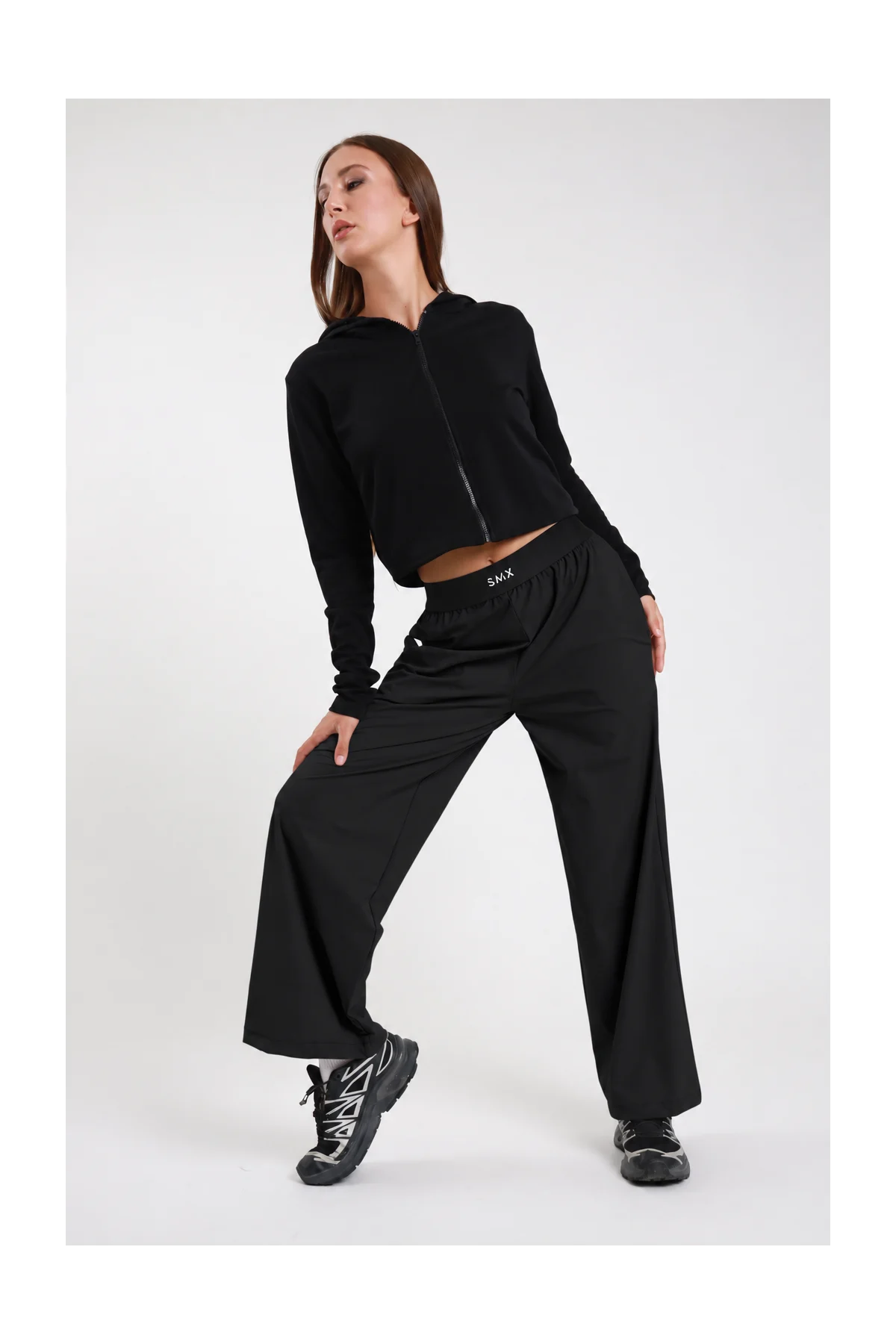 Pantaloni Wide-Leg Soft Air DAYTHRU nero elastico SMX bianco a contrasto gamba wide extra-ampia fluida piega frontale silhouette palazzo lunga frontale intera posa dinamica gamba alzata sguardo basso - modella frontale intera posa dinamica gamba alzata sguardo basso capelli castani lisci sfondo bianco felpa zip crop nera sneakers nere chunky