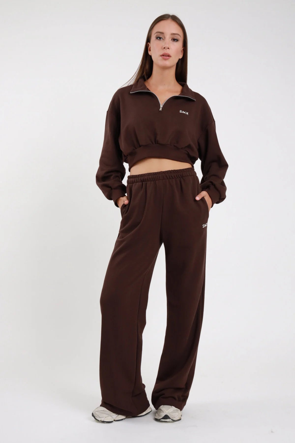 Pantalone Wide Leg DAYTHRU marrone cioccolato scuro elastico vita logo SMX bianco coscia destra gamba wide jogger tasche laterali frontale intera posa statica mani tasche sguardo frontale - modella frontale intera posa statica mani tasche sguardo frontale capelli castani lisci sfondo bianco felpa crop 1/2 zip marrone coordinata sneakers bianche