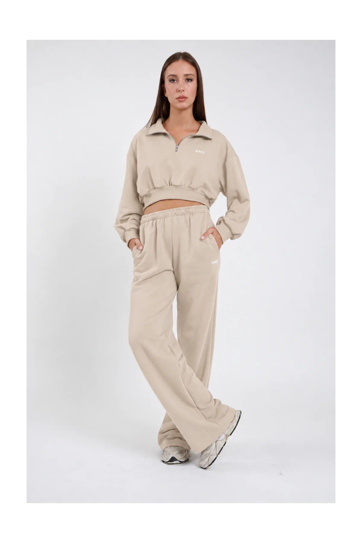 Pantalone Wide Leg DAYTHRU sabbia beige chiaro elastico vita logo SMX bianco coscia gamba wide jogger tasche laterali frontale intera posa statica mano fianco sguardo basso - modella frontale intera posa statica mano fianco sguardo basso capelli castani lisci sfondo bianco felpa crop 1/2 zip sabbia coordinata sneakers grigie
