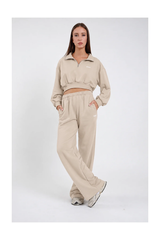 Pantalone Wide Leg DAYTHRU sabbia beige chiaro elastico vita logo SMX bianco coscia gamba wide jogger tasche laterali frontale intera posa statica mano fianco sguardo basso - modella frontale intera posa statica mano fianco sguardo basso capelli castani lisci sfondo bianco felpa crop 1/2 zip sabbia coordinata sneakers grigie