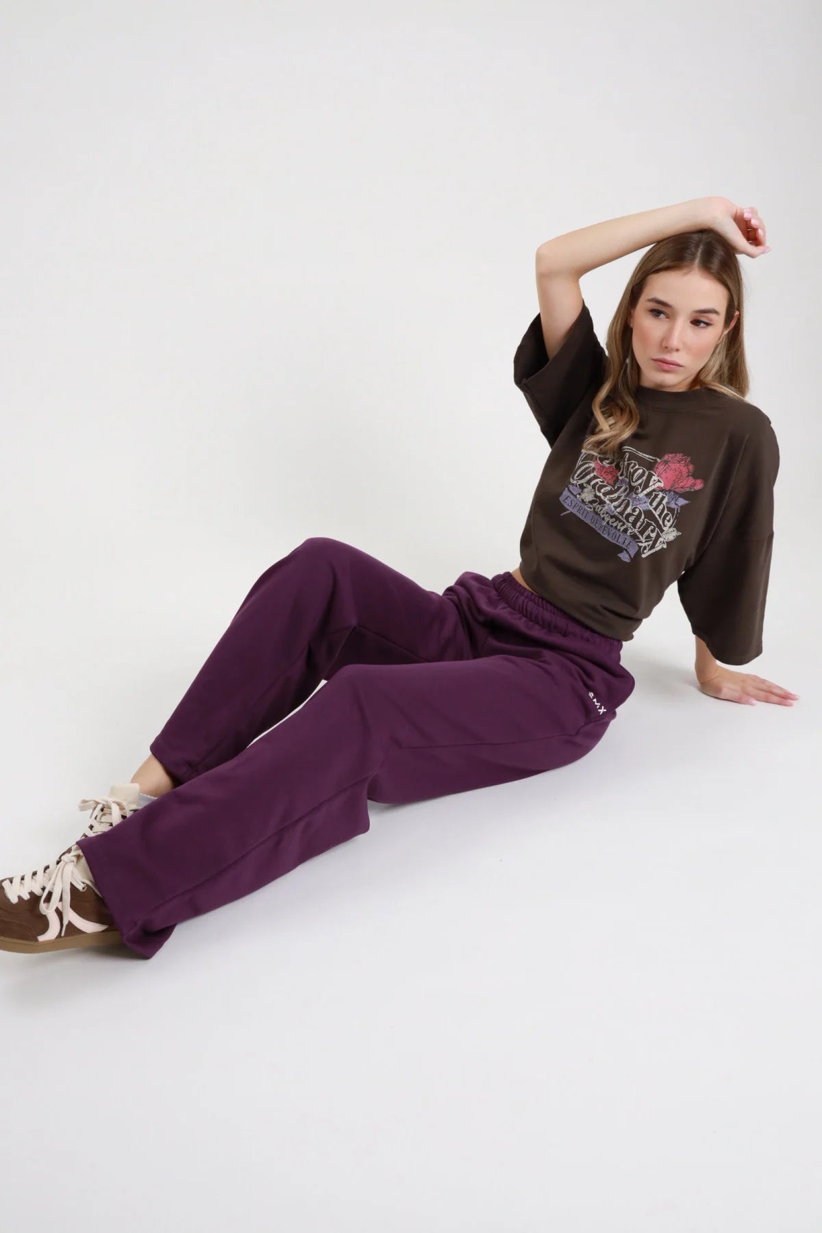 Pantalone Wide Leg SMX Limited Edition viola melanzana vita elastica logo SMX coscia gamba wide leg palazzo polsino fondo arricciato seduta pavimento braccia gomiti gambe laterali - modella seduta pavimento braccia gomiti gambe laterali sguardo frontale capelli castani sfondo bianco sneakers marrone tan t-shirt crop cioccolato grafica abbinata