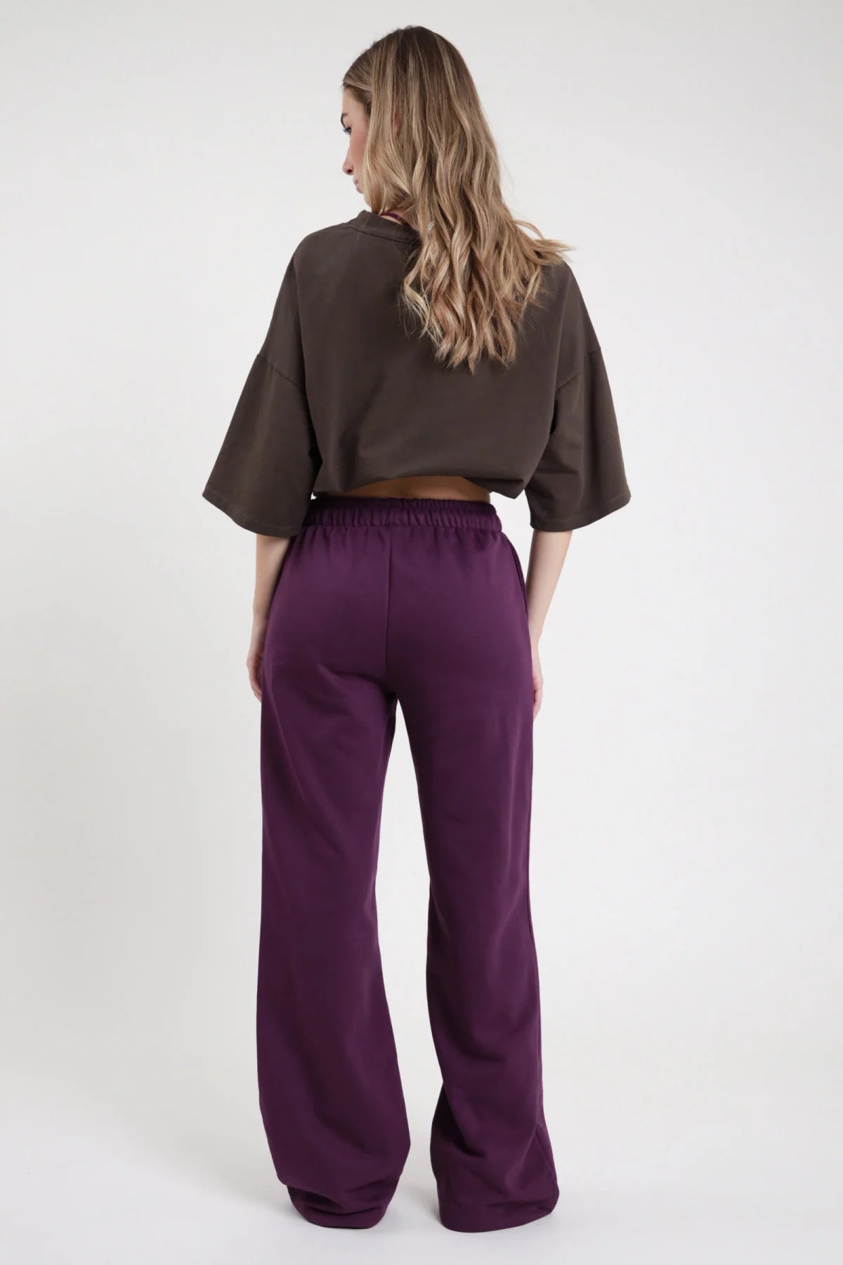 Pantalone Wide Leg SMX Limited Edition viola melanzana 65% cotone french terry vita elastica posteriore gamba wide leg palazzo lungo retro liscio - modella intera posteriore capelli mossi castani chiari sfondo bianco t-shirt crop cioccolato grafica abbinata