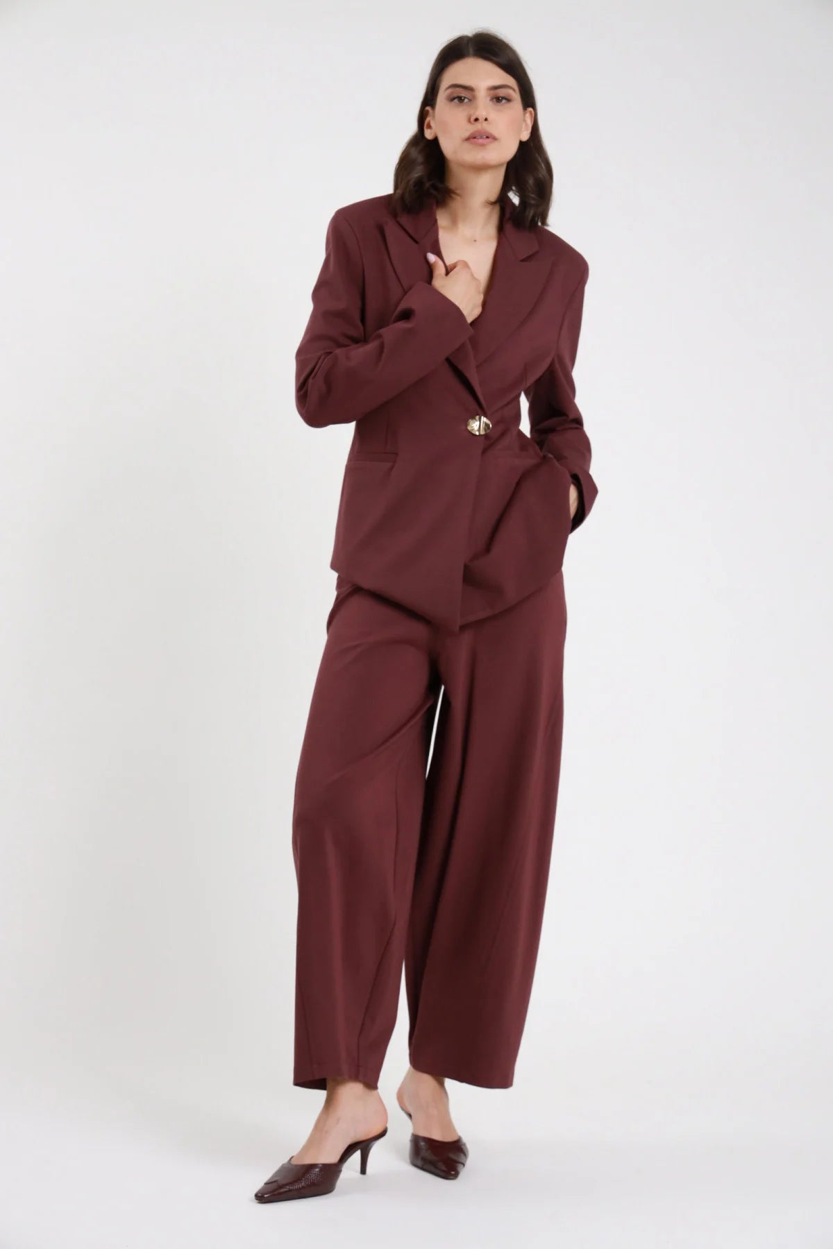 Pantalone Uovo Punto Milano bordeaux vinaccia vita alta pinces gamba barrel scultorea cropped 7/8 frontale intera con blazer bordeaux bottone dorato - modella frontale intera mano colletto sguardo frontale capelli castani scuri sfondo bianco blazer bordeaux bottone dorato mules bordeaux tacco