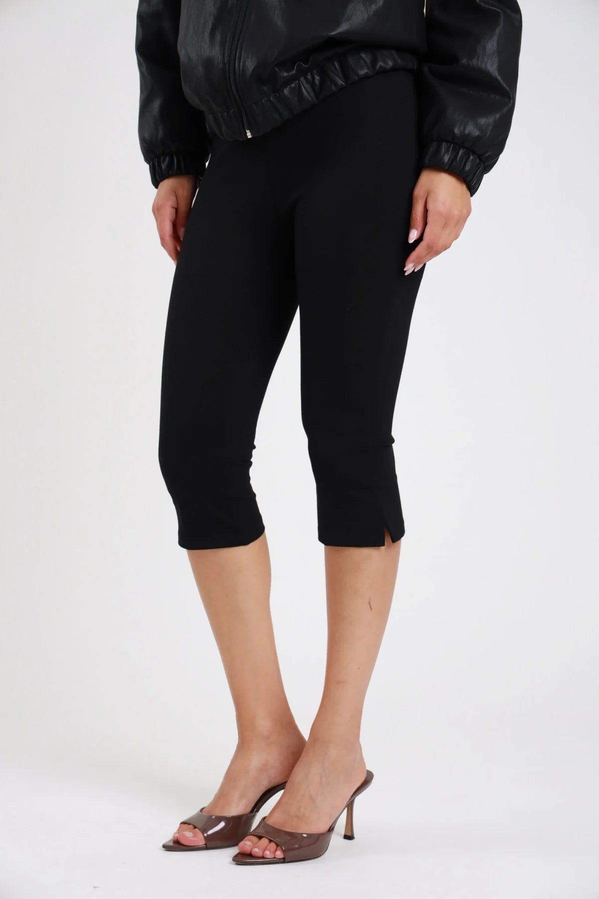 Pantaloni capri pinocchietto nero donna ultra-slim vita alta elasticizzata lunghezza capri spacco laterale - dettaglio frontale tre quarti mani sui fianchi con bomber ecopelle nero e sandali tacco marrone