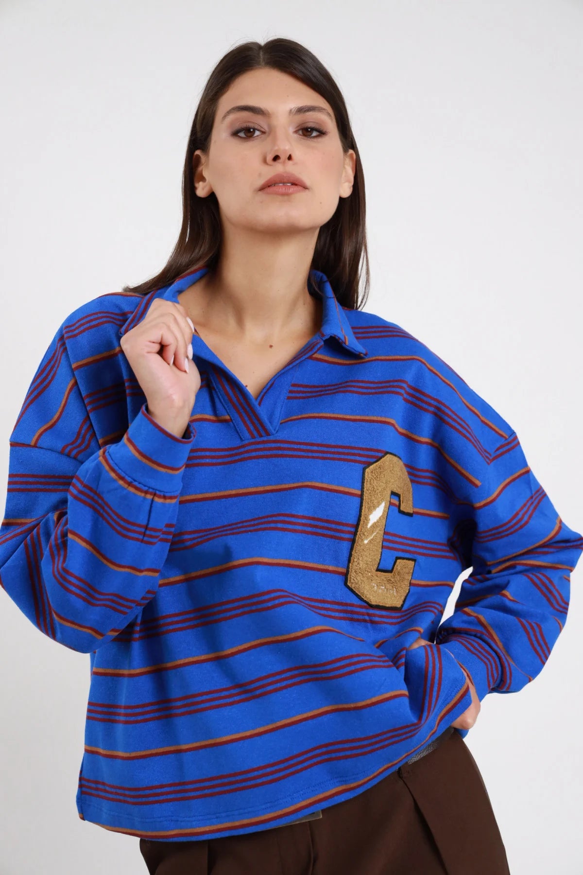 Polo Rugby Oversize Righe Multicolor Patch Varsity blu cobalto elettrico rosso arancio ocra 77% cotone colletto polo aperto V profondo maxi patch lettera C varsity dorata trapuntata petto sx manica ampia drop polsino costina boxy crop frontale - modella busto 3/4 frontale posa mano colletto sguardo frontale intenso make-up bold capelli castani sfondo bianco pantaloni marrone cioccolato