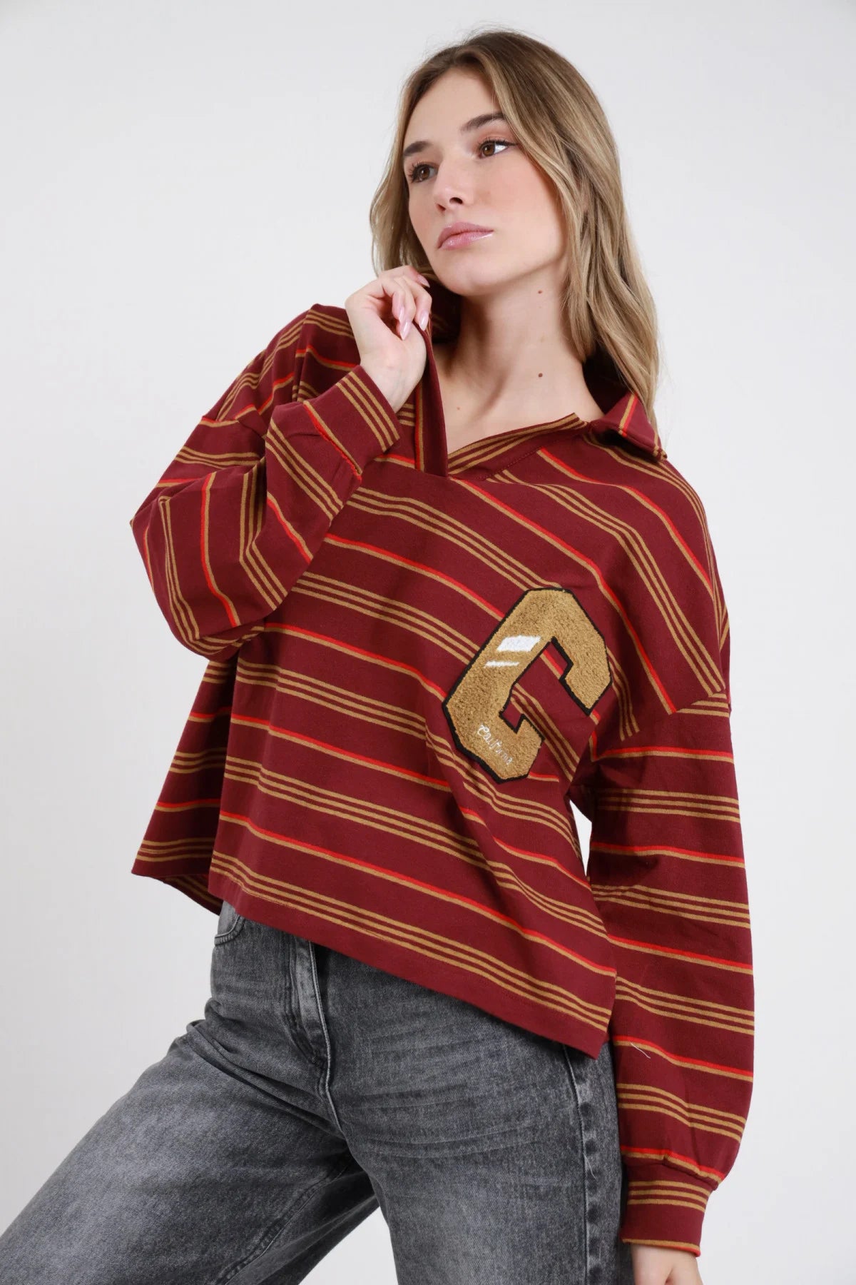 Polo Rugby Oversize Righe Multicolor Patch Varsity bordeaux vinaccia rosso arancio senape ocra 77% cotone colletto polo aperto V maxi patch lettera C varsity dorata trapuntata petto sx manica ampia drop polsino costina boxy crop 3/4 frontale - modella busto 3/4 frontale posa mano colletto sguardo 3/4 soft capelli biondi mossi sfondo bianco jeans grigio antracite