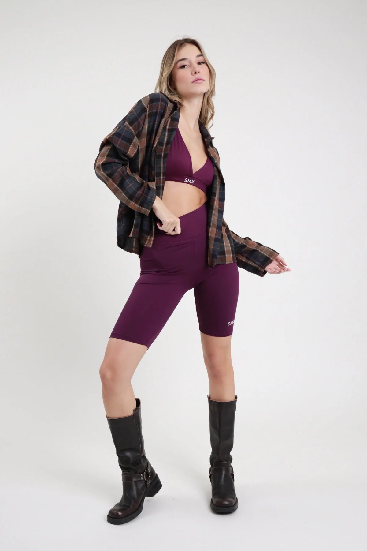 Short Ciclista SMX Limited Edition viola melanzana 95% tactel vita elastica alta banda logo SMX bianco bordo superiore logo coscia laterale lunghezza ginocchio body-fit frontale intera in piedi - modella intera frontale in piedi capelli biondi mossi sfondo bianco bralette triangolo viola SMX coordinata giacca tartan scozzese oversize aperta stivali neri alti ginocchio abbinati