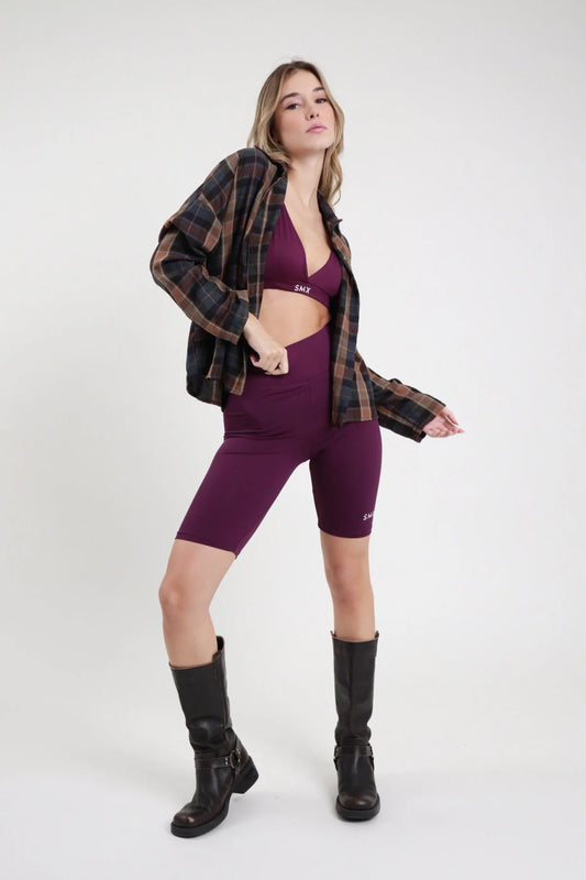 Short Ciclista SMX Limited Edition viola melanzana 95% tactel vita elastica alta banda logo SMX bianco bordo superiore logo coscia laterale lunghezza ginocchio body-fit frontale intera in piedi - modella intera frontale in piedi capelli biondi mossi sfondo bianco bralette triangolo viola SMX coordinata giacca tartan scozzese oversize aperta stivali neri alti ginocchio abbinati