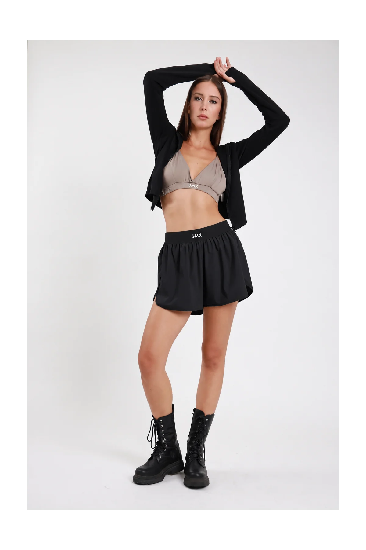 Short Soft Air Skort DAYTHRU nero elastico nero logo SMX tono su tono gonna sovrapposta arricciata fluida ampia silhouette skort frontale intera posa dinamica braccia alzate testa sguardo frontale - modella frontale intera posa dinamica braccia alzate testa sguardo frontale capelli castani lisci sfondo bianco top triangolo taupe SMX felpa crop zip nera aperta stivali combat neri