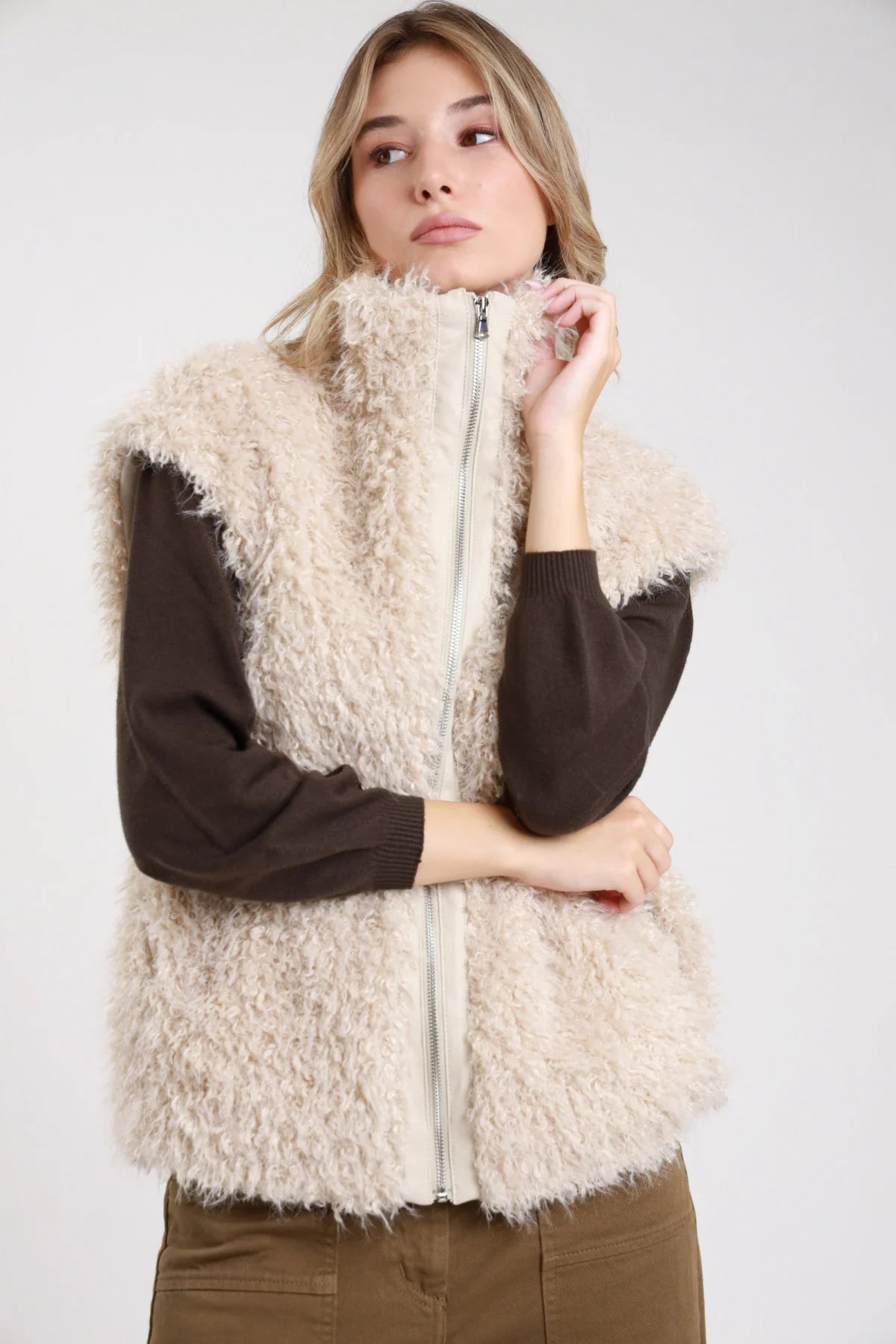 Gilet ecopelliccia riccioluta sabbia panna colletto alto zip argento - modella busto frontale mano al colletto con maglia cioccolato e jeans kaki capelli biondi raccolti