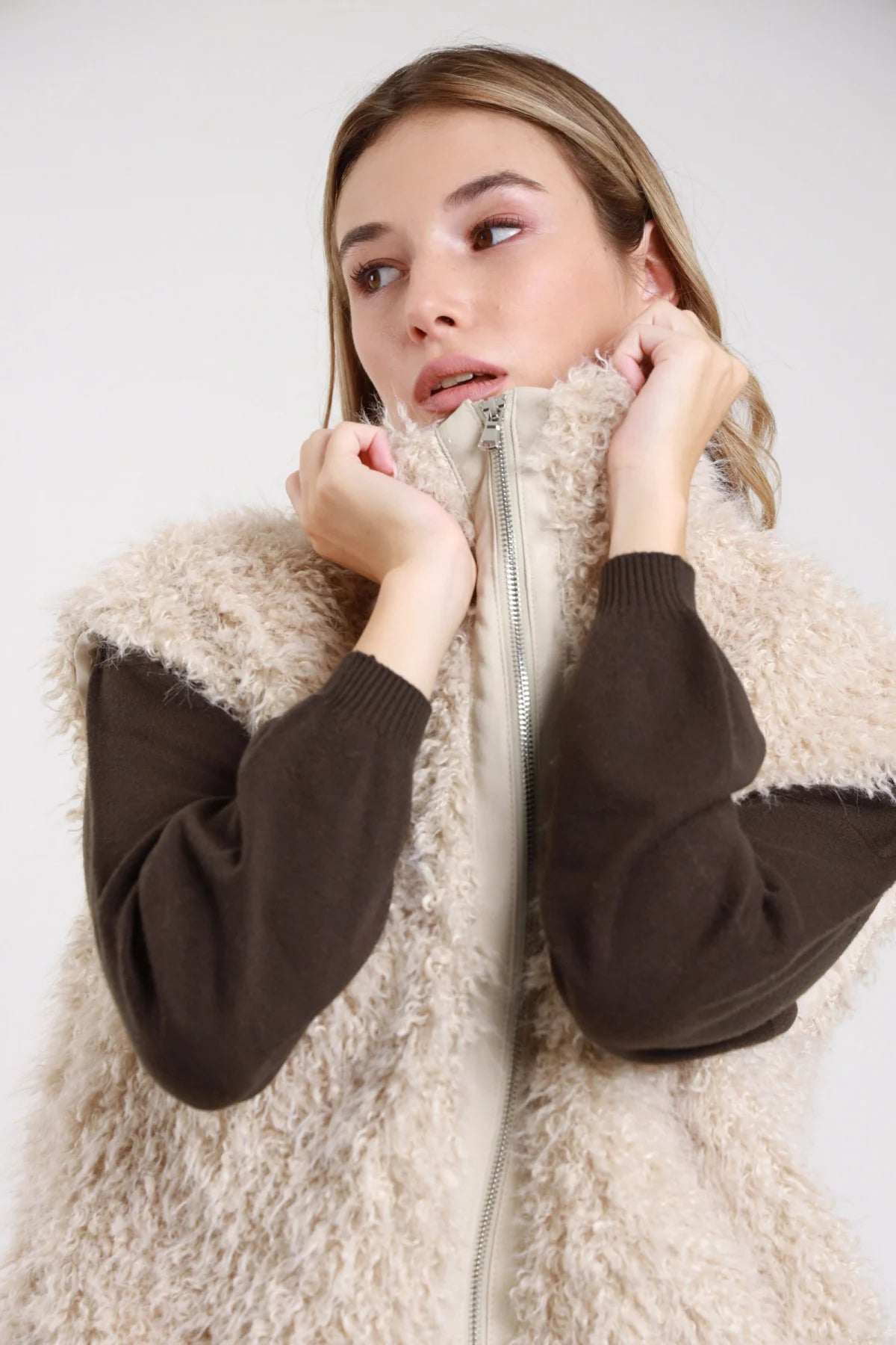 Gilet ecopelliccia shearling teddy bear zip aperta dettaglio colletto - modella busto ravvicinato mani che tengono colletto con maglia cioccolato sguardo laterale capelli biondi