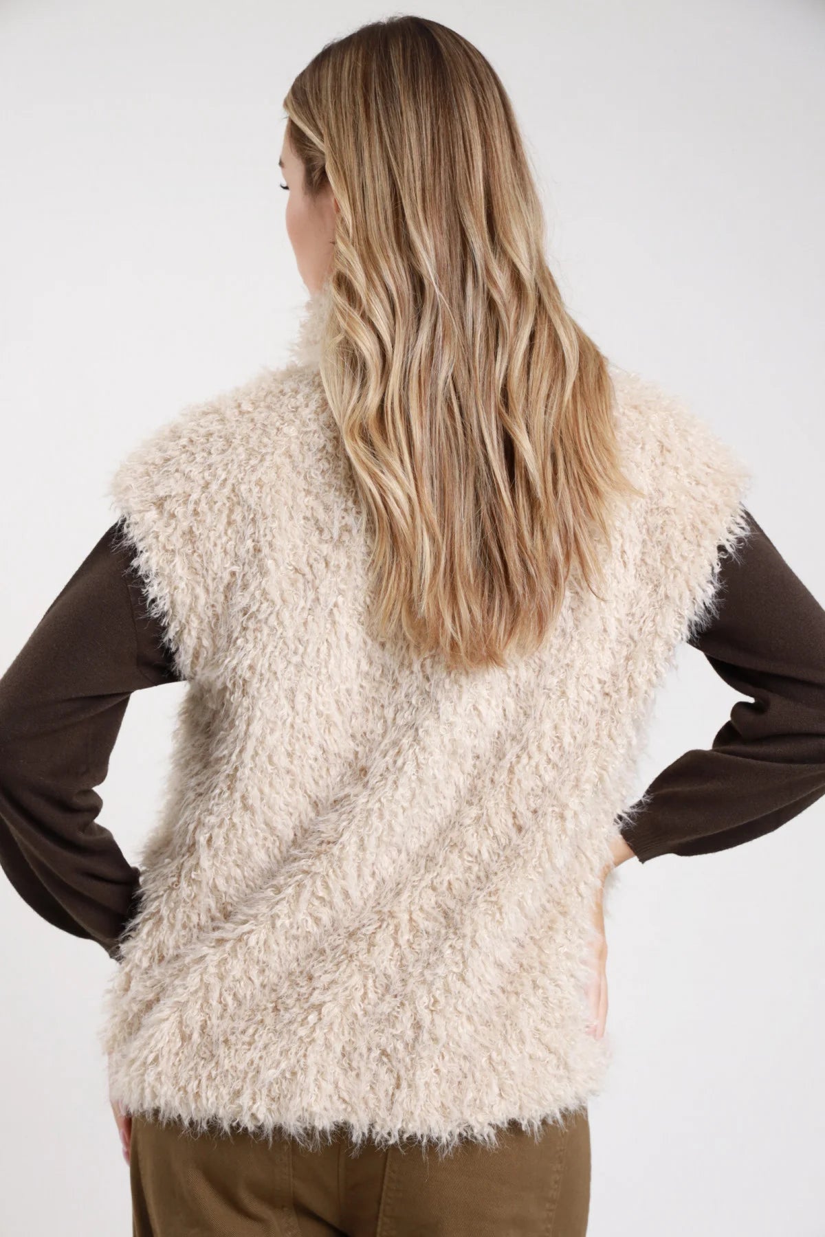 Gilet ecopelliccia riccioluta sabbia panna boxy dritto - modella retro intera mani sui fianchi con maglia cioccolato jeans kaki capelli biondi sciolti