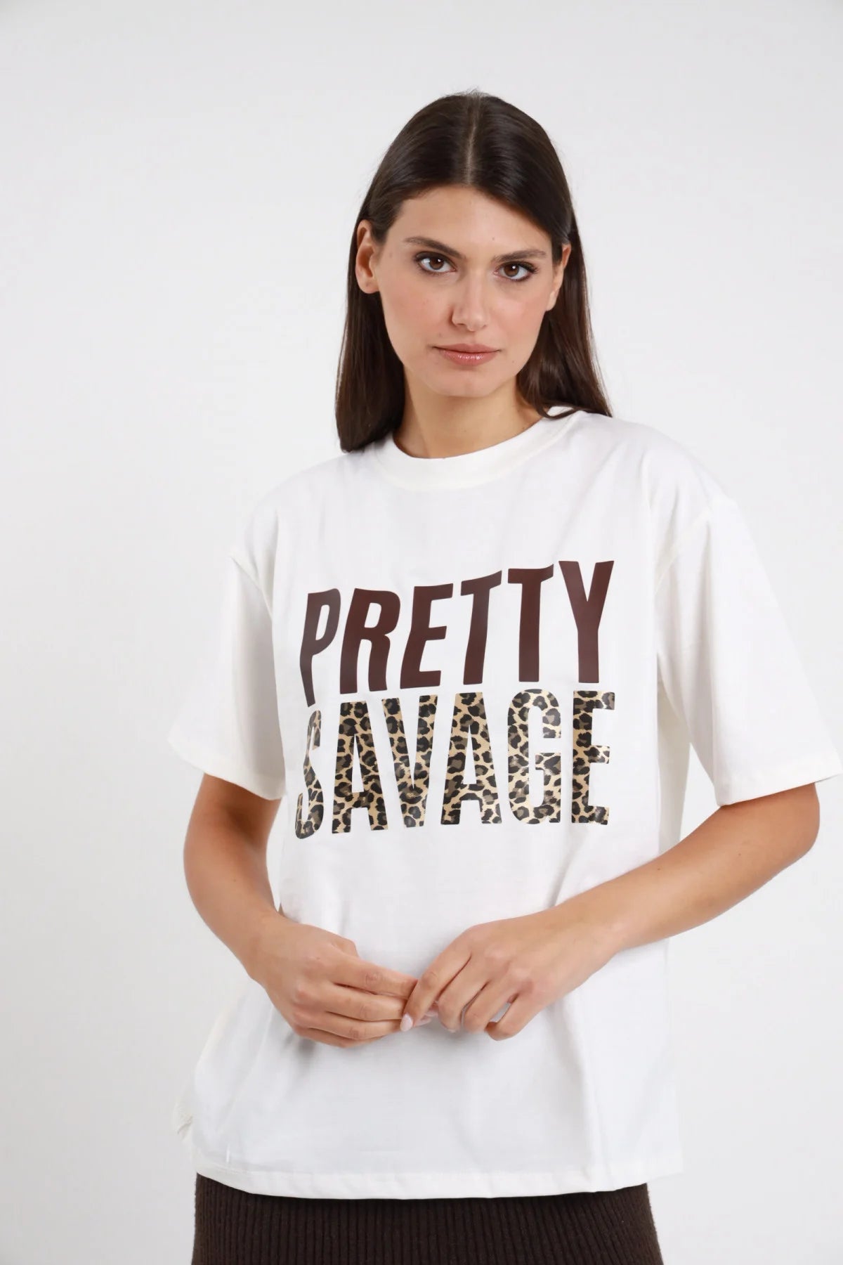 T-shirt Pretty Savage bianco panna 100% cotone stampa PRETTY marrone scuro SAVAGE leopardato marrone beige girocollo manica corta silhouette oversize - modella busto frontale posa mani grembo sguardo frontale intenso capelli castani lisci sfondo bianco gonna nera a coste
