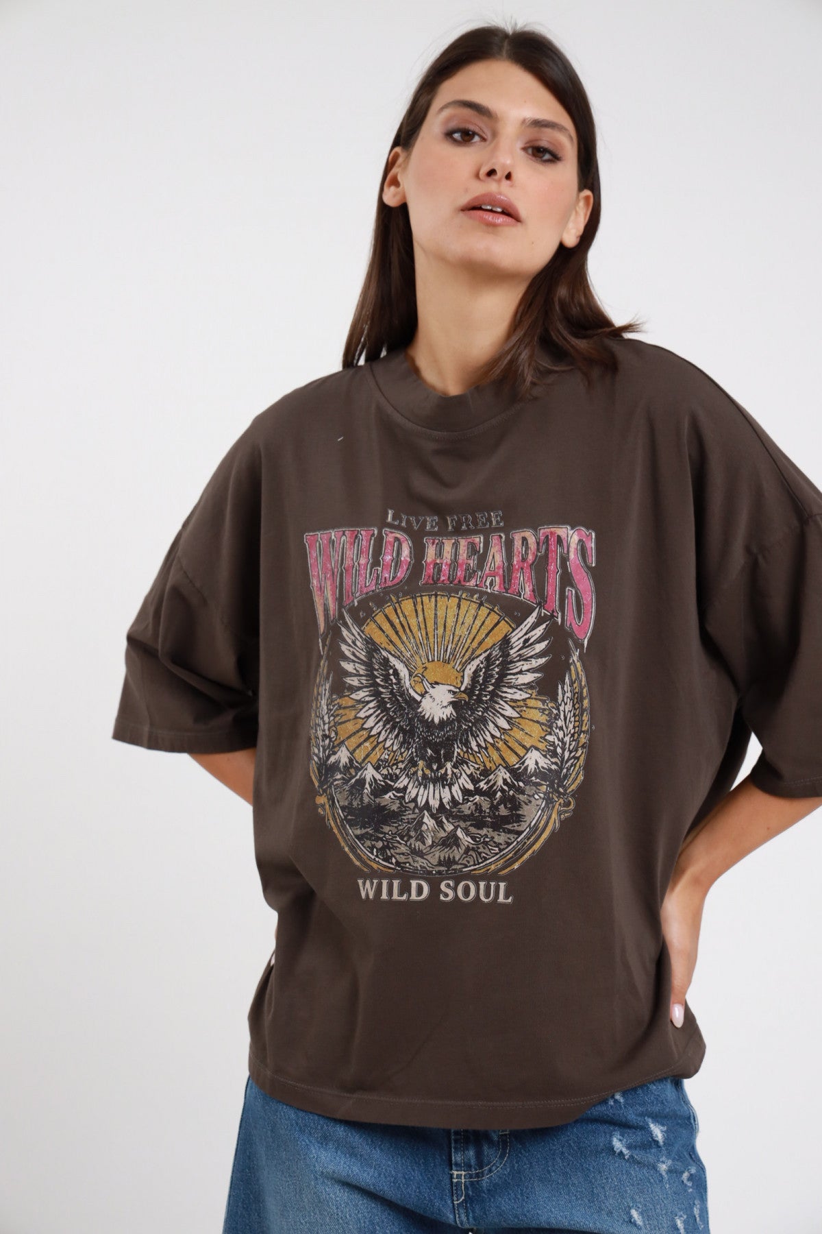 T-shirt Aquila cioccolato marrone scuro 95% cotone oversize boxy girocollo drop shoulder stampa grafica aquila americana Wild Hearts Wild Soul vintage western frontale busto 3/4 - modella busto 3/4 sguardo frontale capelli castani scuri lisci sfondo bianco jeans distressed strappati abbinati