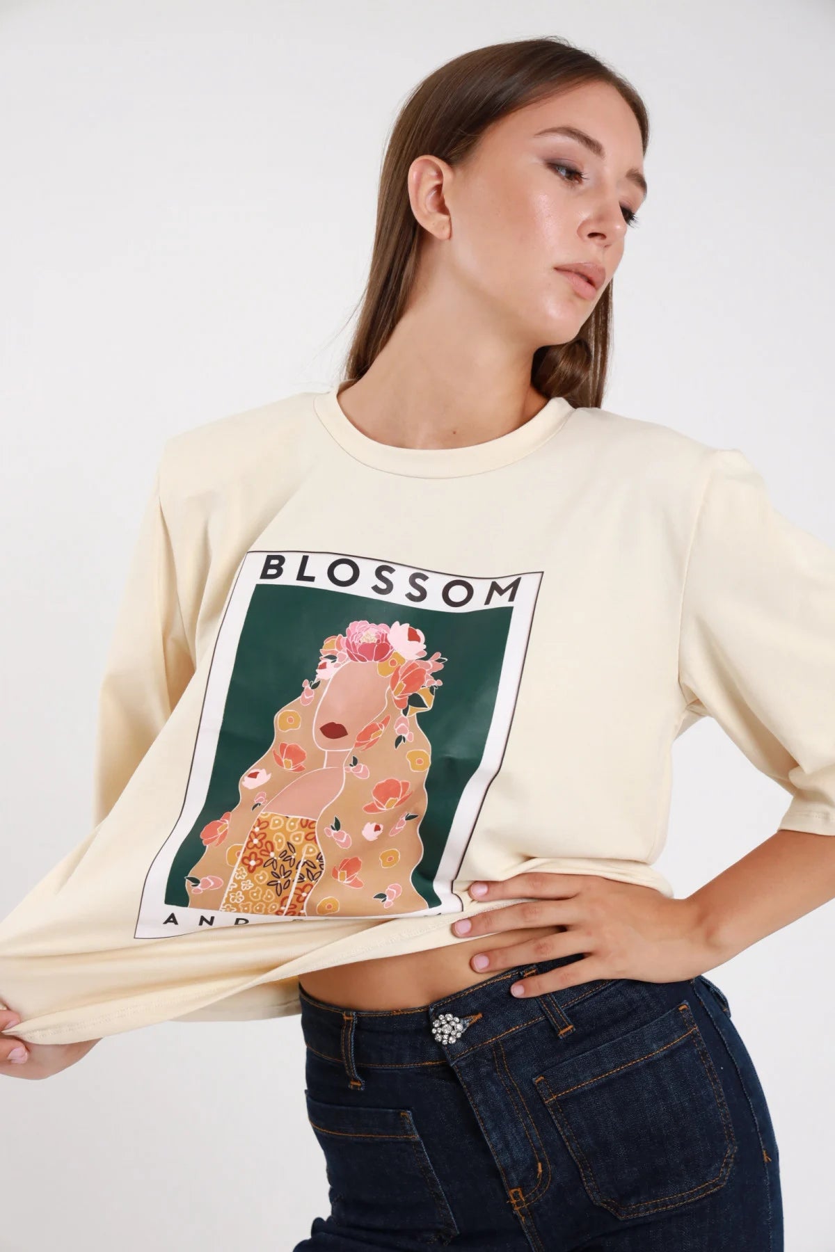 T-shirt blossom bloom panna ecru busto tre quarti stampa art nouveau klimt blossom bloom cornice bianca sollevata - modella busto tre quarti mano lembo t-shirt sguardo laterale alto jeans scuro capelli castani raccolti