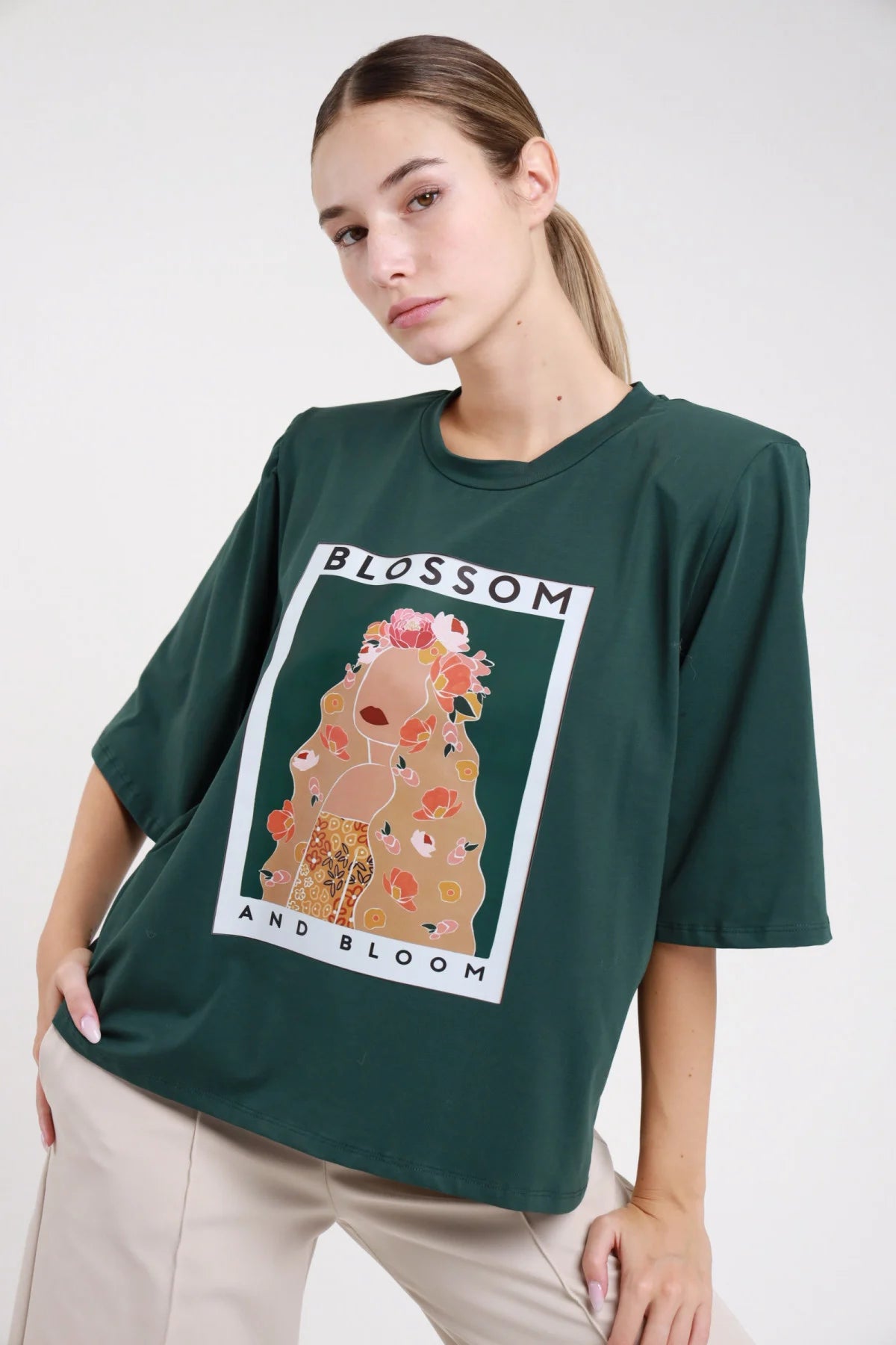 T-shirt blossom bloom verde bottiglia frontale busto girocollo stampa art print figura femminile fiorita cornice polaroid - modella frontale busto mano fianco sguardo frontale pantaloni beige capelli castani raccolti