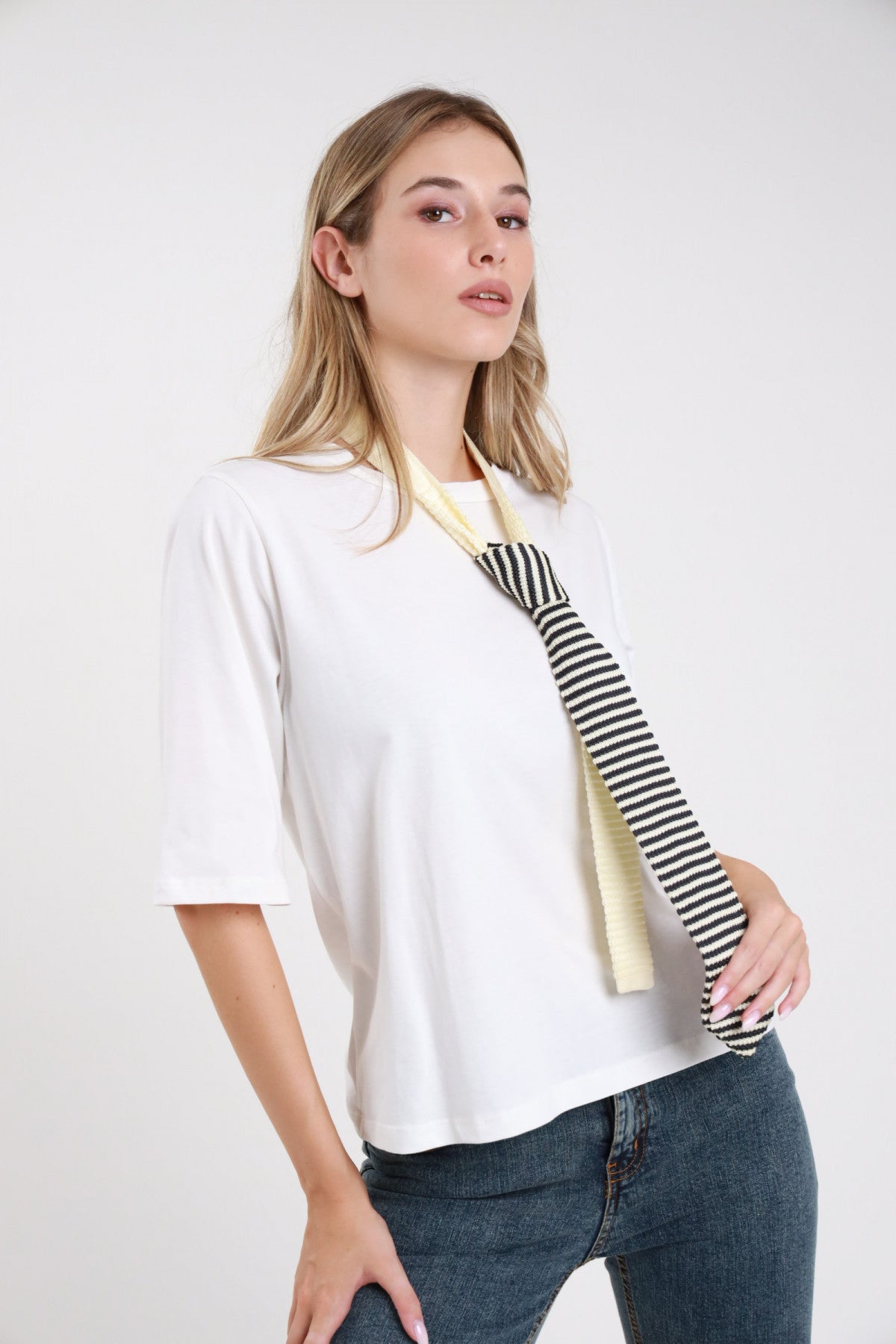 T-shirt bianca oversize con cravatta maglia righe bordeaux giallo panna colletto raso cioccolato girocollo manica corta dropped shoulder - modella frontale busto mano cravatta sguardo laterale basso jeans moro capelli castani raccolti