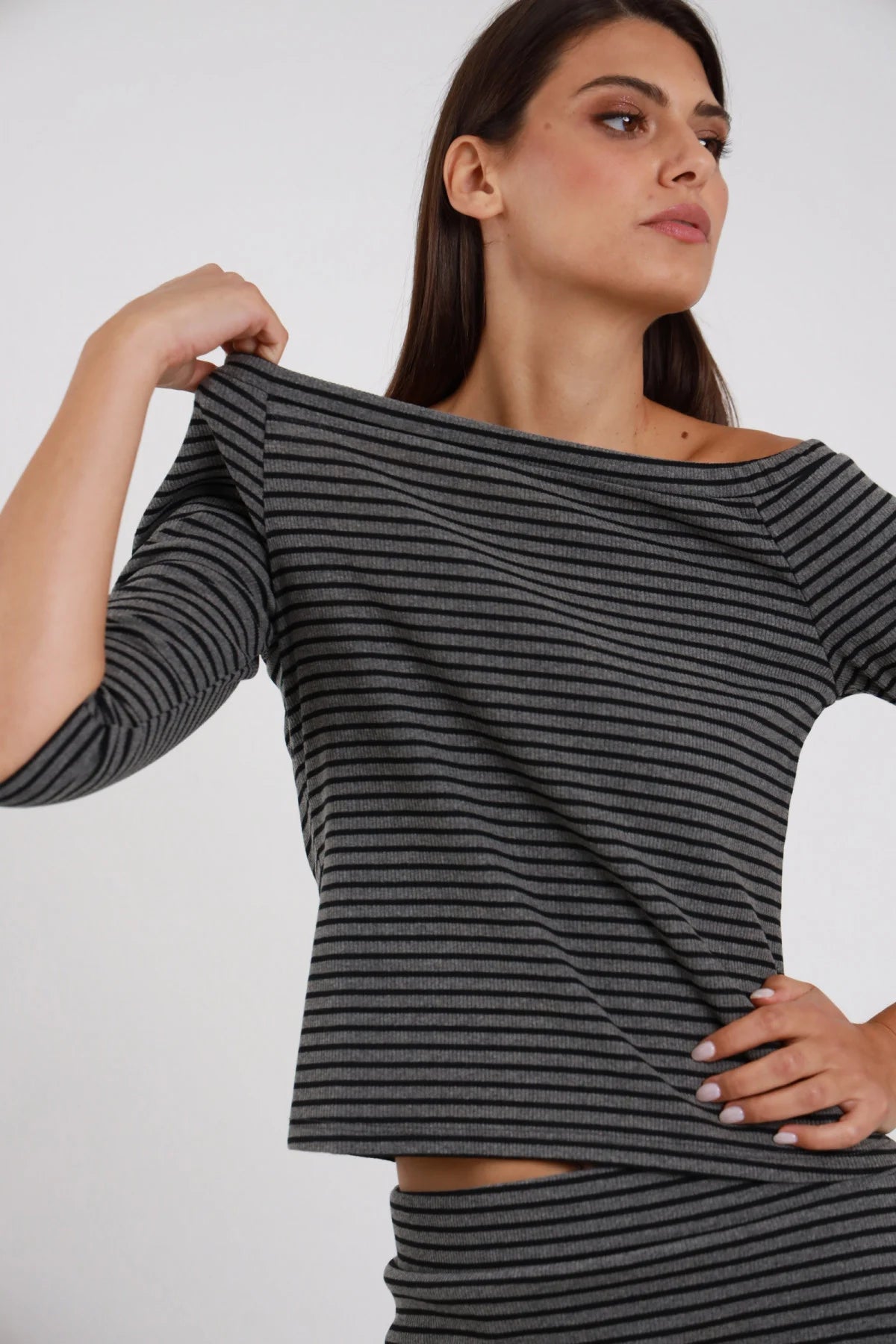 Top Righe Lurex grigio antracite nero 48% cotone scollo barchetta ampio off-shoulder manica 3/4 silhouette crop semi-aderente - modella busto 3/4 frontale posa mano tira colletto barchetta lateralmente sguardo laterale alto capelli castani lisci sfondo bianco gonna coordinata righe grigie