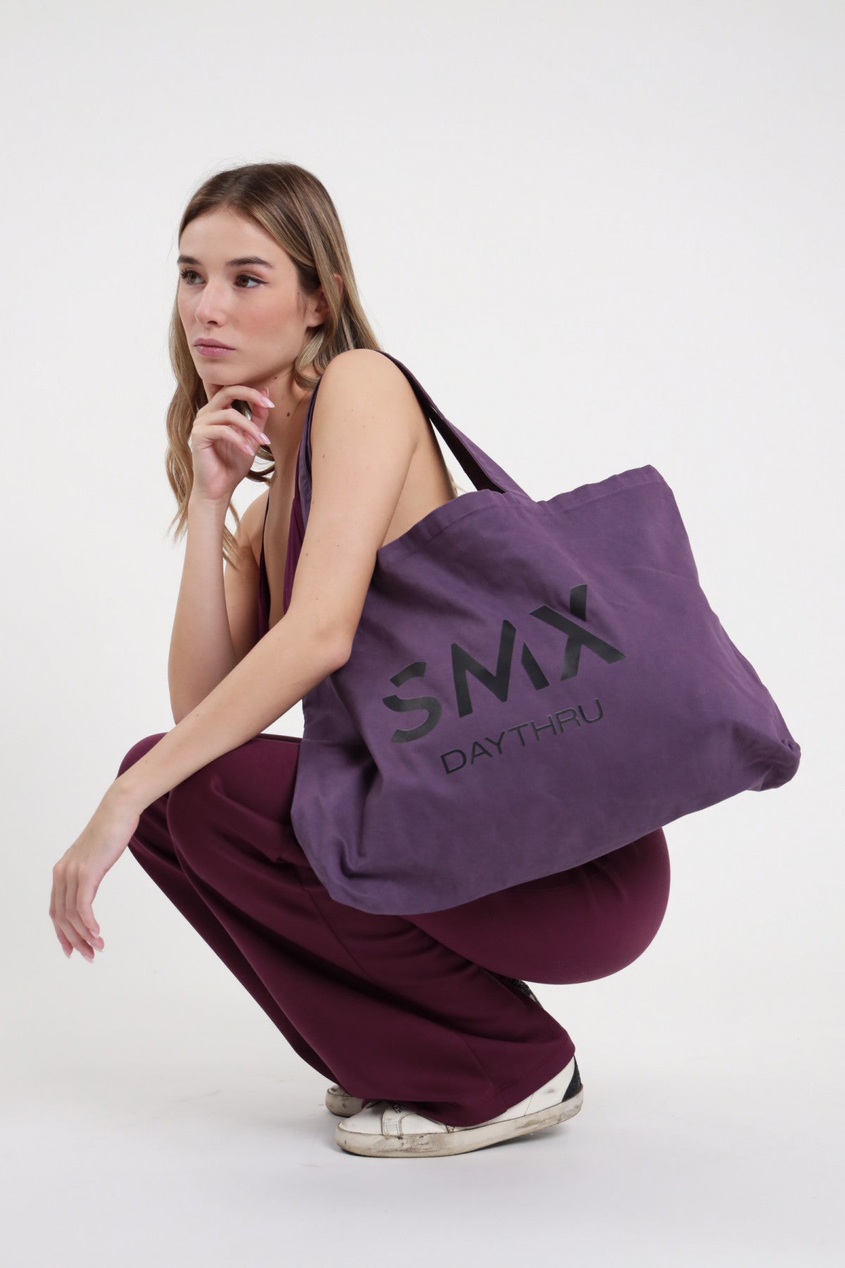 Tote Bag SMX Daythru Limited Edition viola melanzana 100% cotone canvas stampa logo SMX DAYTHRU nero tono su tono manici lunghi spalla maxi shopper capiente - modella accovacciata laterale mano mento sguardo laterale capelli biondi mossi sfondo bianco top bordeaux senza spalline pantalone wide leg bordeaux flare sneakers bianche abbinati