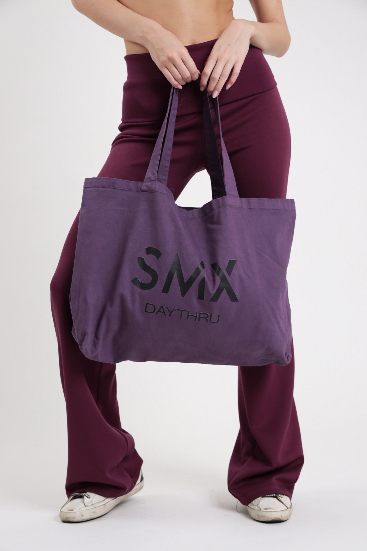 Tote Bag SMX Daythru Limited Edition viola melanzana 100% cotone canvas logo SMX DAYTHRU frontale manici lunghi mani manico gambe larghe - modella frontale mani manici gambe larghe sfondo bianco pantalone wide leg bordeaux flare sneakers bianche abbinati