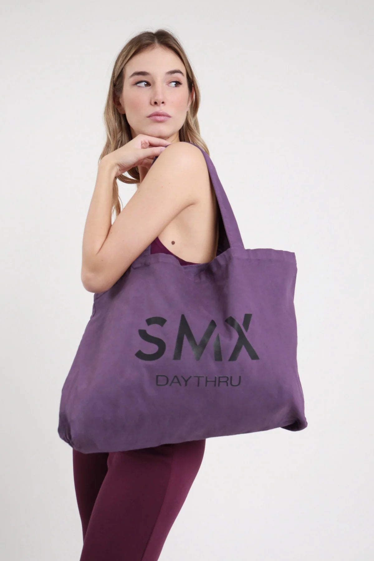 Tote Bag SMX Daythru Limited Edition viola melanzana prugna 100% cotone canvas stampa logo SMX DAYTHRU nero tono su tono manici lunghi tessuto spalla formato maxi shopper - modella busto 3/4 laterale spalla mano mento sguardo alto capelli biondi mossi sfondo bianco top bordeaux senza spalline leggings bordeaux abbinati