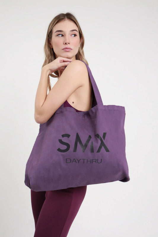 Tote Bag SMX Daythru Limited Edition viola melanzana prugna 100% cotone canvas stampa logo SMX DAYTHRU nero tono su tono manici lunghi tessuto spalla formato maxi shopper - modella busto 3/4 laterale spalla mano mento sguardo alto capelli biondi mossi sfondo bianco top bordeaux senza spalline leggings bordeaux abbinati