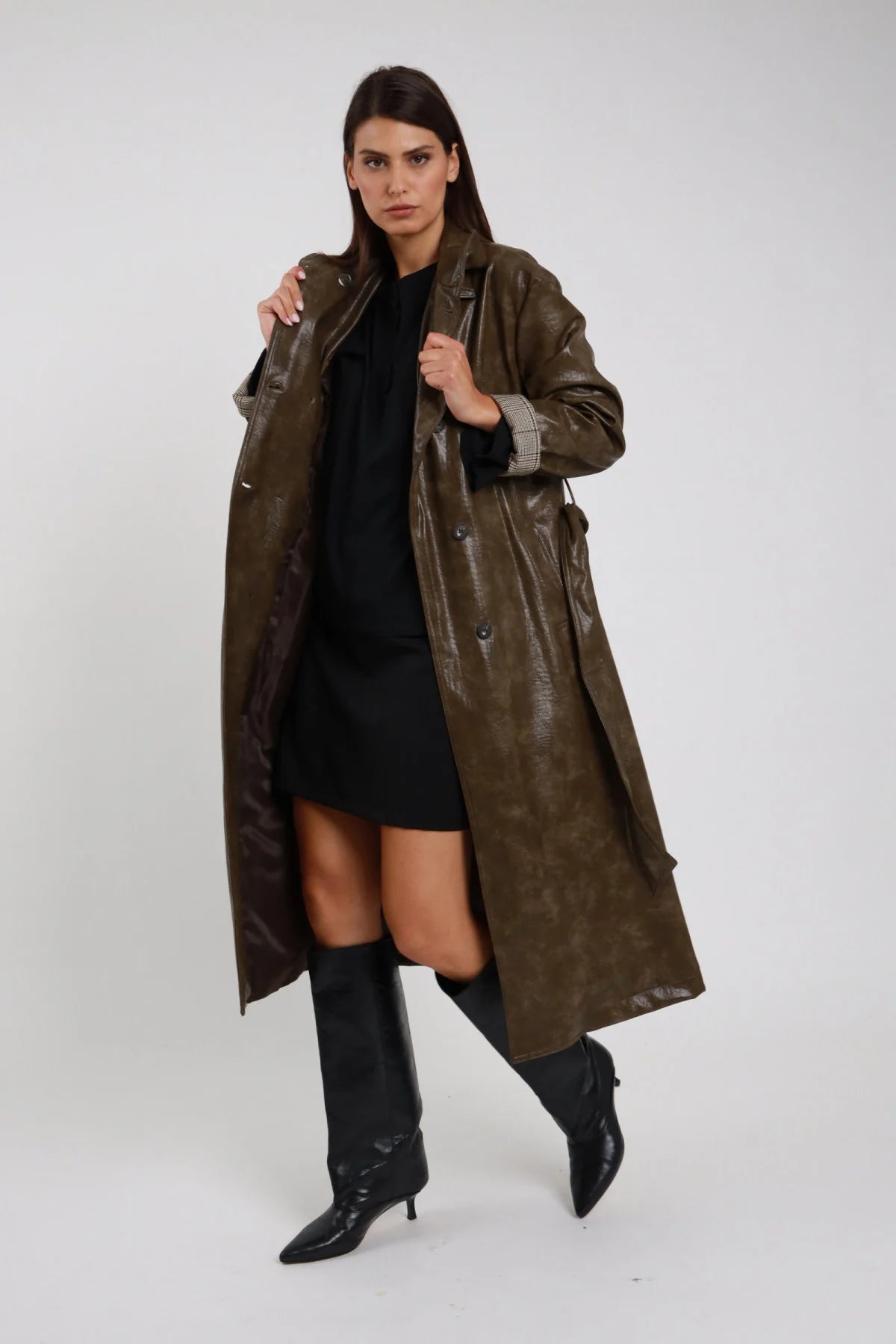 Trench Ecopelle verde oliva kaki scuro verniciata laccata crinkle colletto camicia doppiopetto tasche frontali polsino check kaki lunghezza maxi midi frontale intera posa aperta mani colletto - modella frontale intera posa aperta mani colletto sguardo laterale capelli castani lisci sfondo bianco vestitino nero stivali neri alti tacco kitten